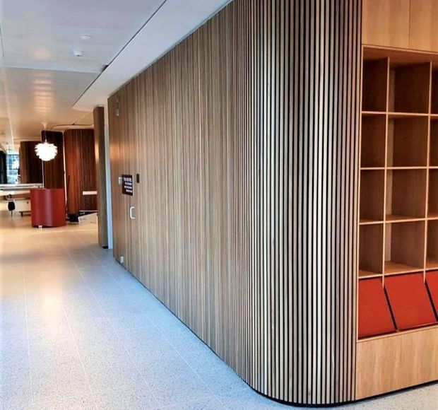 Interior view of a contemporary commercial lobby or corridor with a curved wall clad in closely spaced vertical timber slats, integrated timber shelving, red upholstered seating, pale tiled flooring, and modern pendant lighting.