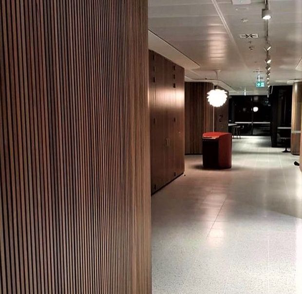 A modern commercial interior corridor or lobby with extensive vertical timber slat wall panelling, integrated timber cabinetry, a pale polished terrazzo or concrete floor, and a suspended track-lighting system. A small red reception or display pedestal and a sculptural pendant light provide accents, while dark glazing and an illuminated exit sign appear in the background.