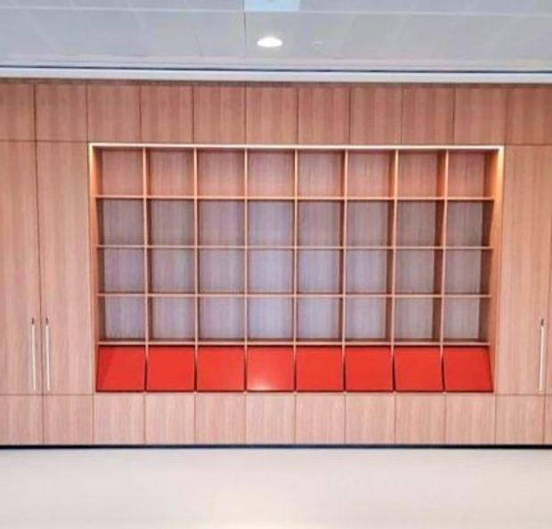 A full-height built-in timber-toned storage and display wall with a large open grid of shelving. The lower shelving row has vivid red drawer or cabinet fronts, while tall closed cabinets flank both sides. The installation is shown in a clean, bright interior beneath a white panelled ceiling with recessed lighting.