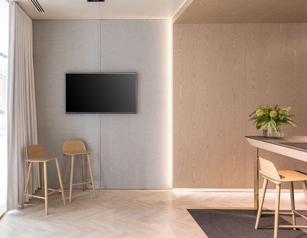 A refined, contemporary interior featuring a wall-mounted television on a pale textured wall, light timber wall panelling and cabinetry, a compact bar or kitchen counter with timber stools, and light herringbone timber flooring. Full-height curtains and glazing brighten the space, while concealed linear lighting and simple furnishings create a clean, warm atmosphere.