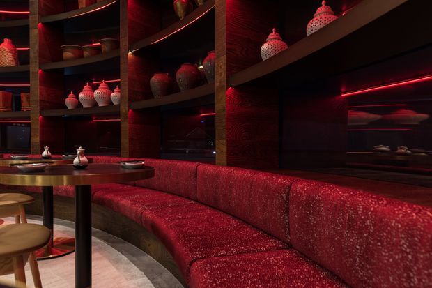 A dramatic, richly layered restaurant or lounge interior featuring a curved red upholstered banquette wrapping around a small round dining table. Dark timber shelving and wall panels display decorative ceramic vessels, while integrated red linear lighting creates a moody, luxurious atmosphere. A large dark reflective surface or pass-through is visible behind the seating.