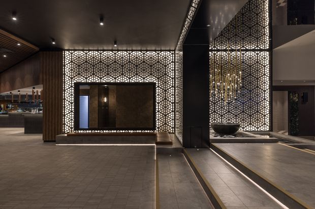 A dramatic, upscale interior lobby or entry space featuring illuminated geometric lattice screens, dark stone or tile flooring, timber wall panelling, a large framed feature opening, stepped platforms, and a reflective water or decorative basin element. Recessed spotlights and a sculptural chandelier create a moody, luxurious atmosphere. A bar or hospitality area is visible in the background.