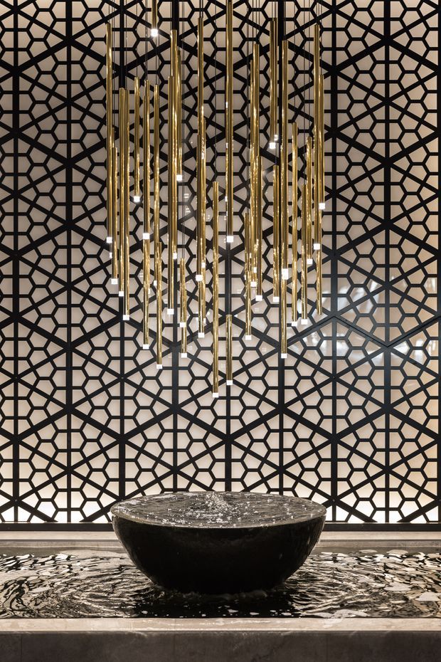 A dramatic, symmetrical interior feature with a black stone bowl fountain set in a shallow reflective water basin. Behind it is a full-height geometric black metal screen with an intricate Islamic-inspired pattern, while numerous suspended brass and glass pendant lights form a sculptural chandelier overhead. The palette is predominantly black, warm metallic gold, and neutral stone, creating a luxurious and refined atmosphere.