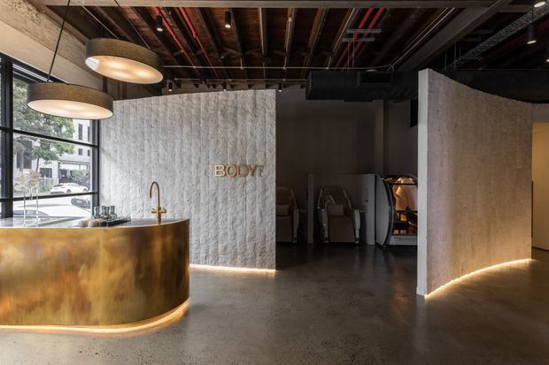 Interior of a dark, industrial-style wellness or fitness studio reception area. A curved metallic reception desk with an integrated sink sits beside large street-facing windows. Textured pale concrete or plaster partitions, exposed timber ceiling structure, visible services, black ductwork, pendant lighting, and concealed floor-level lighting define the space. A dark doorway leads toward treatment or exercise areas, with a small illuminated BODY sign on the partition.