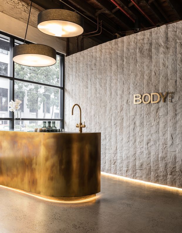 An industrial-style reception or service counter featuring a curved brushed brass front, integrated brass tap, and warm underlighting. The space has exposed dark ceiling structure and services, large black-framed windows, a textured pale stone or concrete block feature wall with illuminated “BODY” signage, and a polished concrete floor.