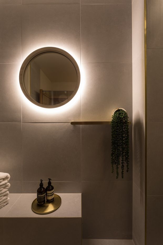 A warmly lit contemporary bathroom vignette featuring large taupe-grey wall tiles, a round backlit mirror, a slim brass shelf with trailing greenery, and a stone or solid-surface ledge holding folded towels and dark soap dispensers on a brass tray. The space has a refined, moody and minimalist atmosphere.