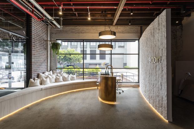 Industrial-style commercial interior with a curved built-in upholstered bench, compact reception or service island, exposed brick and concrete surfaces, visible ceiling services, large black-framed windows, and warm linear accent lighting. A textured feature wall with signage defines the right side of the space.