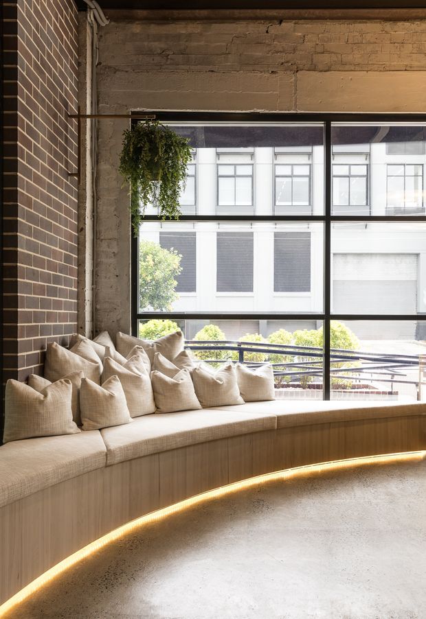 A warm, contemporary commercial-style lounge or waiting area with a curved built-in timber banquette upholstered in neutral fabric and layered cushions. Exposed brick walls, a rough concrete ceiling and floor, black-framed industrial glazing, and concealed amber under-seat lighting create a refined industrial atmosphere. A hanging plant and an urban building are visible through the large window.