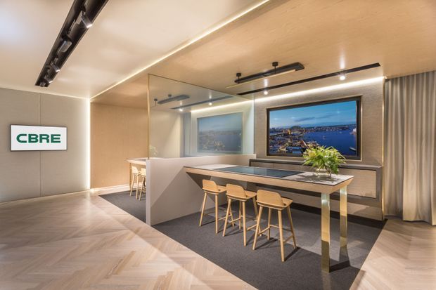 A refined contemporary commercial hospitality or meeting lounge with a large central island table and four timber bar stools. The space features warm wood-look ceiling and herringbone flooring, charcoal carpet, bronze-toned metal detailing, integrated linear and track lighting, full-height curtains, and mirrored wall panels. Framed city and waterfront imagery is displayed on the rear wall, and a CBRE sign is visible on the left.