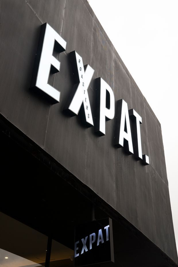 Low-angle exterior view of a contemporary commercial building facade clad in dark panels, featuring large projecting white EX/PAT lettering and a hanging illuminated EX/PAT sign above the entrance.