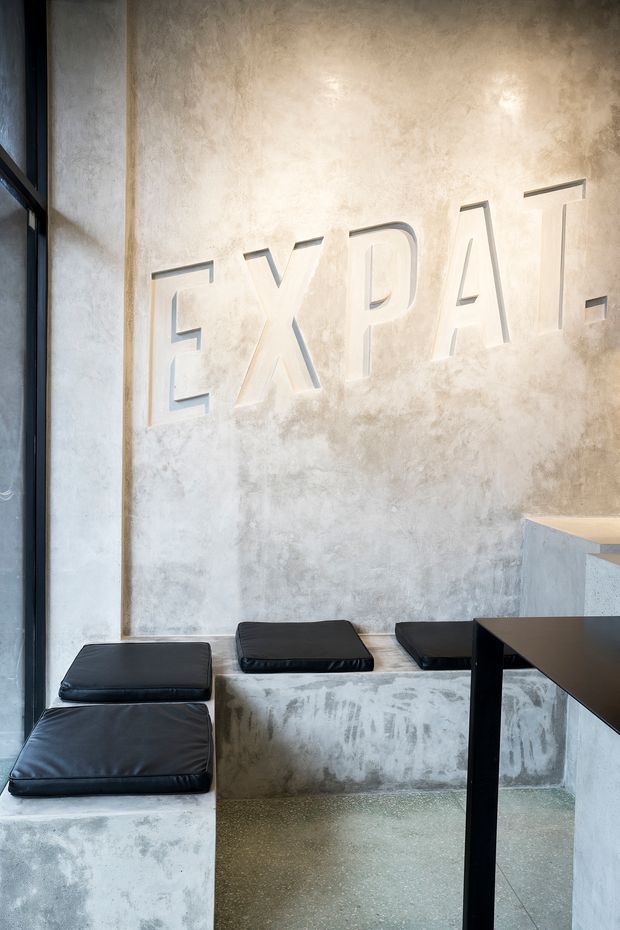 A minimalist commercial interior featuring a sculptural concrete or plaster seating bench with black leather cushions, a dark tabletop, and large raised lettering on a textured pale wall. Tall black-framed glazing brings in natural light, while the space uses a restrained grey and black palette with raw, industrial finishes.