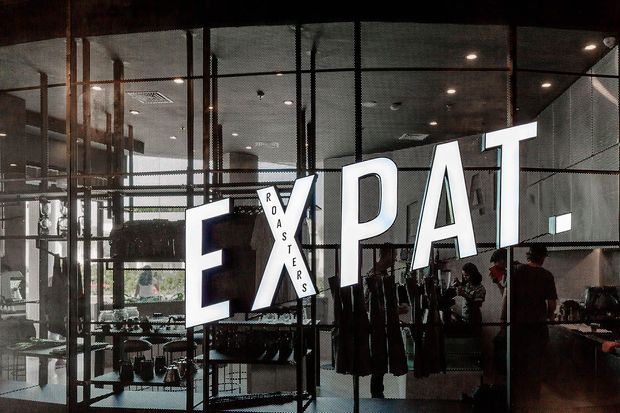 Interior view of a contemporary retail showroom or commercial space with extensive black-framed glass partitions, exposed dark ceiling structure, display shelving, clothing racks, and large illuminated 'EXPAT ROASTERS' signage. The space has an industrial, warehouse-like character with visible activity and merchandise displays.