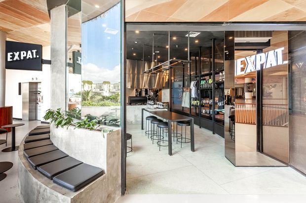 A bright contemporary commercial interior, likely a café or bar, with exposed concrete columns, polished light flooring, black-framed glass partitions, dark cabinetry, a central bar counter with stools, and built-in curved bench seating by full-height glazing. Black, timber, and metallic finishes create a refined industrial atmosphere.