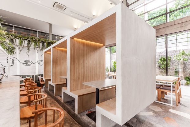 A bright contemporary commercial dining space with a series of timber-lined booth seating bays framed by white architectural partitions. Warm integrated lighting highlights the natural timber joinery, while timber dining chairs and tables sit on a patterned hard floor. Large industrial-style glazed windows, exposed concrete elements, hanging greenery, and wall artwork contribute to the open café or restaurant atmosphere.
