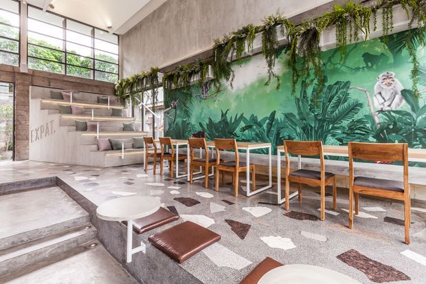 A bright, high-ceilinged hospitality-style interior with a long communal dining table and timber chairs arranged against a dramatic tropical mural. Hanging greenery runs along the upper wall, while large industrial-style windows, polished concrete surfaces, terrazzo-like flooring, built-in stepped seating, and a small round table create a contemporary café or restaurant atmosphere.