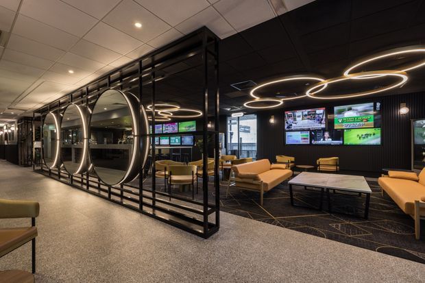 A sophisticated commercial lounge or hospitality space with a dark, contemporary interior. A black metal partition with large circular decorative openings separates the seating area from the corridor. The room features tan leather lounge chairs and sofas, low tables, multiple wall-mounted screens, black vertical wall panelling, patterned dark carpet, and distinctive illuminated ring-shaped ceiling lights.