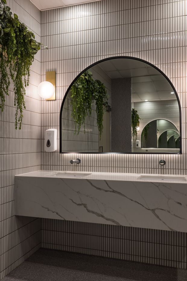 A refined contemporary bathroom vanity area featuring a large floating double vanity with a white marble-look surface and pronounced grey veining, slim vertically ribbed wall tiles, a large black-framed arched mirror, spherical wall lighting, and cascading greenery. The dark floor and warm lighting create a sophisticated, slightly moody atmosphere.