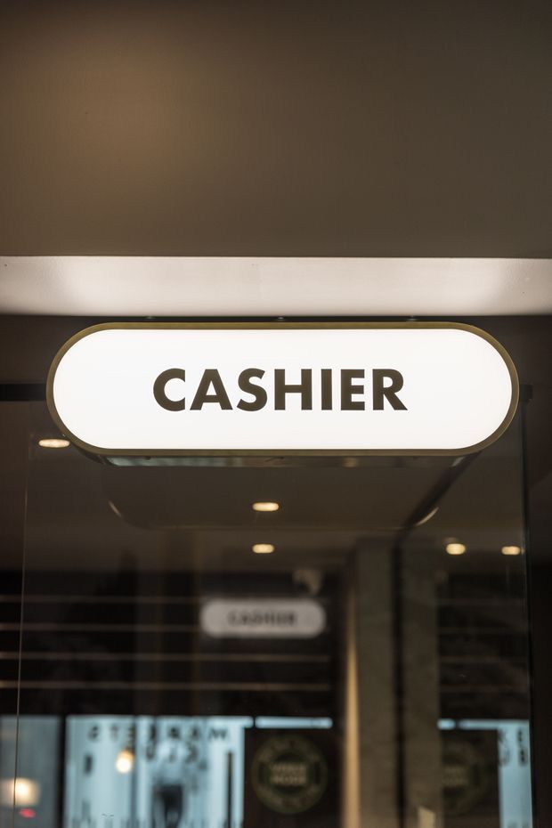 Close-up photograph of a backlit, rounded rectangular “CASHIER” sign mounted above a reflective glazed storefront or counter area. The sign has dark lettering, a warm metallic-looking border, and is set within a dim, modern commercial interior with visible ceiling lights and reflections.