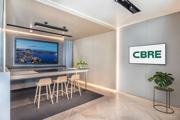Contemporary commercial reception or breakout lounge with a compact high table and four timber stools, wall-mounted display screen, CBRE branding, integrated linear and track lighting, pale timber herringbone flooring, light wood ceiling, and textured grey wall panels. A small indoor plant adds greenery to the polished, professional workspace.