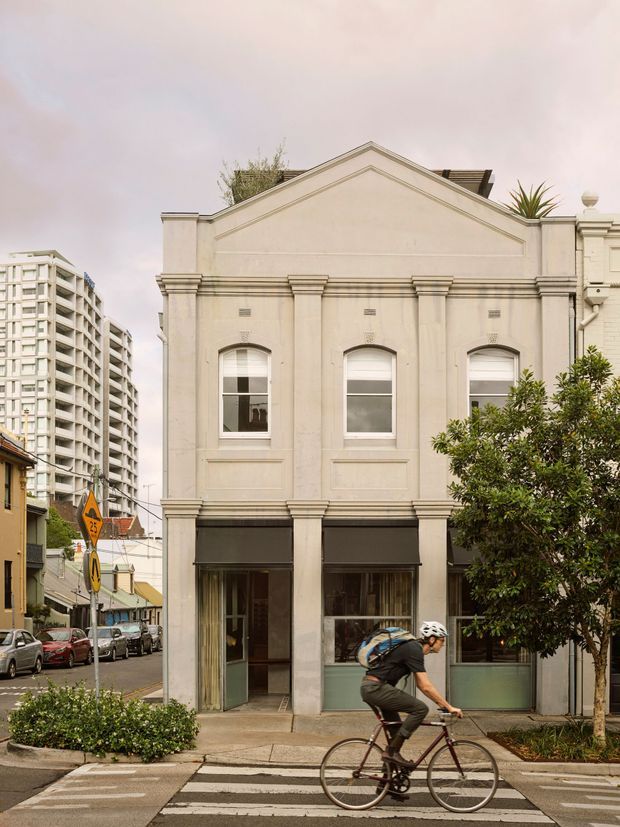 Street-level view of a narrow, restored cream-coloured heritage-style building with a symmetrical classical facade, pediment, pilasters, cornices, arched upper windows, and ground-floor shopfront glazing. A cyclist crosses in front, with an urban streetscape and a taller apartment building visible nearby.