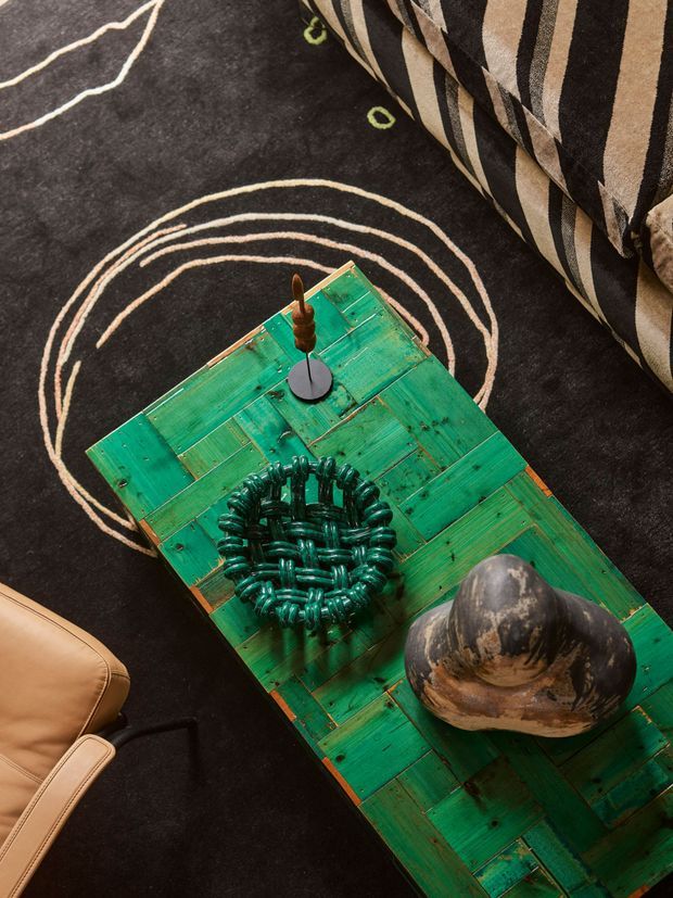 Close-up overhead view of a styled living-room arrangement. A distressed green timber coffee table with visible plank construction and patina holds sculptural decorative objects, including a dark green woven or coiled vessel and a large dark organic-form ornament. The table sits on a black rug with cream abstract curved lines, bordered by a tan leather chair and a black-and-cream patterned upholstered sofa or cushion. The composition has a bold, eclectic, tactile character.