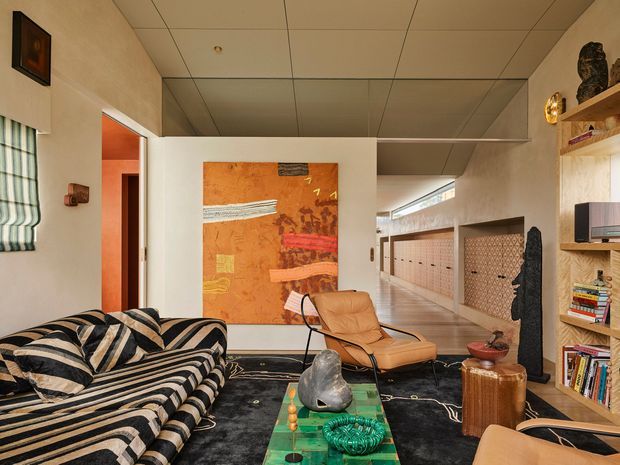 A richly styled mid-century-inspired living room with a bold black-and-cream striped sofa, tan leather lounge chair, dark patterned rug, and a green glass coffee table. Built-in timber shelving displays books and decor, while a large abstract artwork and sculptural accents add colour. The room opens toward a tiled passage or adjoining area, with warm orange tones visible through a doorway and a mirrored or reflective ceiling contributing visual depth.