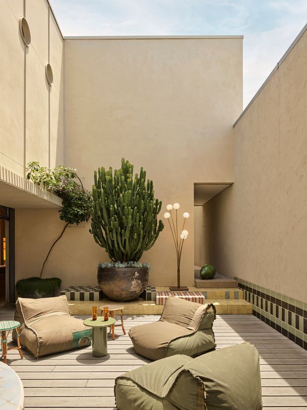 A sunlit, enclosed courtyard terrace with tall warm beige rendered walls and a timber deck. The space features large relaxed floor cushions, small side tables, a tall potted cactus, climbing greenery, patterned tiled steps and a sculptural floor lamp. The architecture is minimalist and contemporary, with a sheltered outdoor living atmosphere and strong natural textures.
