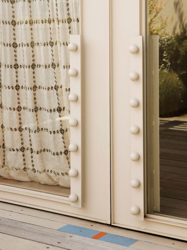 Close-up architectural detail of a cream-painted glazed door or window assembly with rows of rounded decorative studs framing the opening. A patterned sheer curtain is visible behind the left pane, while the right pane opens toward a timber deck and garden. The image emphasizes decorative joinery and warm, refined detailing.