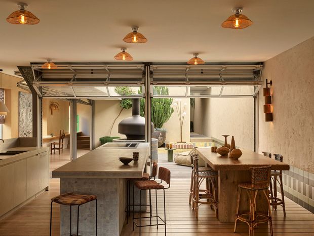 A warm, open-plan kitchen and dining area with a large stone island, timber dining table, woven bar stools, and industrial-style pendant lights. Exposed steel framing and glazed sectional garage-style doors open toward a planted courtyard or patio, while a dark suspended fireplace and textured plaster walls add character.