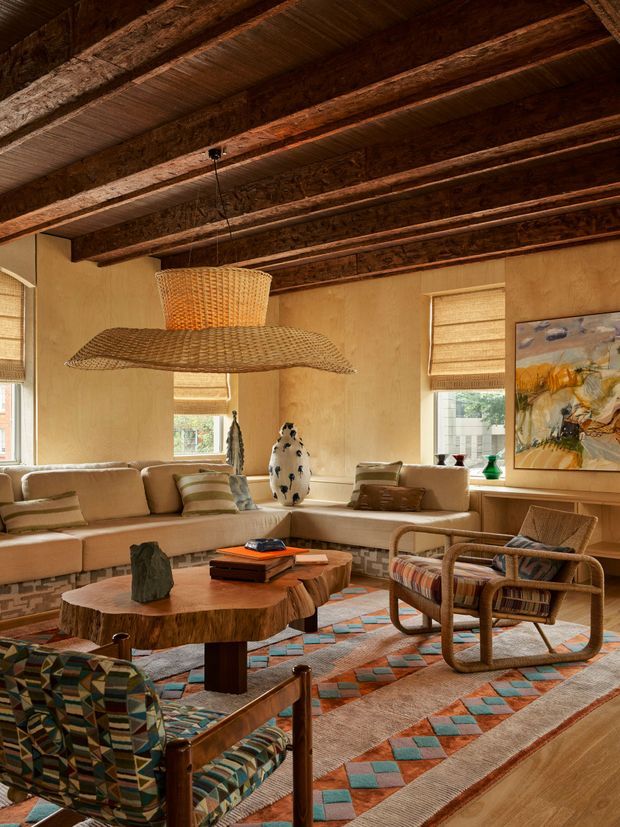Warm, richly layered living room with exposed dark timber ceiling beams, textured tan plaster walls, woven pendant lighting, Roman shades, a large sectional sofa, sculptural wood coffee tables, wicker and patterned lounge chairs, and a colourful geometric rug. The room has an eclectic, earthy, handcrafted character with abundant natural materials and warm daylight.