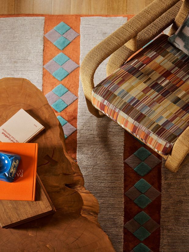 Top-down view of a warmly styled sitting area featuring a woven rattan armchair with a multicoloured geometric patterned cushion, a sculptural live-edge timber coffee table, and layered striped and diamond-patterned rugs. The palette combines terracotta orange, brown, cream, teal, muted pink, and mustard tones, creating a richly textured, retro-inspired interior.
