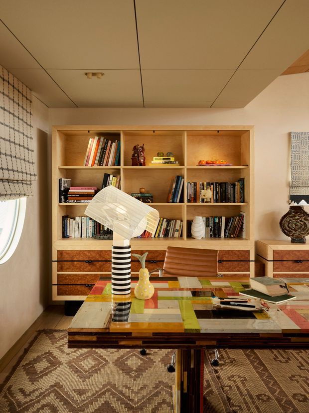 A richly styled home office with a large built-in timber bookcase and drawers occupying the rear wall. The shelves hold books and decorative objects, while a desk in the foreground features a colourful geometric patchwork surface, a striped table lamp, stationery, books, and a sculptural vase. A leather desk chair, patterned rug, textured wall coverings, warm neutral walls, and a wood-lined ceiling create an eclectic, warm, highly decorative interior.
