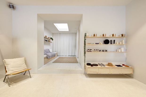 Minimalist, softly lit white interior with a built-in open shelving display and low timber cabinetry holding ceramics and small objects. A recessed, slightly raised sitting or sleeping nook is visible through a wide opening, with sheer curtains, a skylight, a low built-in bench or bed, and a neutral rug. The foreground includes a simple timber-framed lounge chair, while pale continuous flooring and walls create a calm, gallery-like atmosphere.