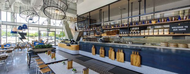 Bright contemporary restaurant or cafe interior with a long navy service counter, open shelving displaying bottles and crockery, hanging wooden cutting boards, patterned tiled counter fronts, exposed ceiling services, large windows, and distinctive black circular pendant lights. Seating and tables occupy the foreground and left side.