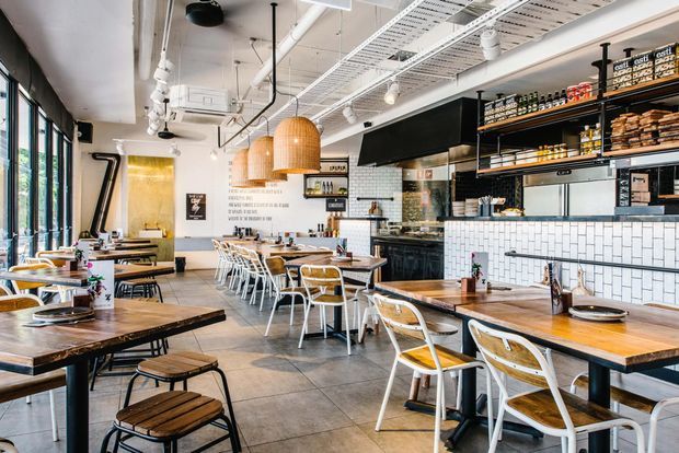 Bright contemporary café or casual restaurant interior with extensive timber dining tables, white and timber chairs, a tiled service counter, open shelving, pendant lighting, and exposed ceiling services. Large windows line one side, while the counter and food-preparation area occupy the rear.