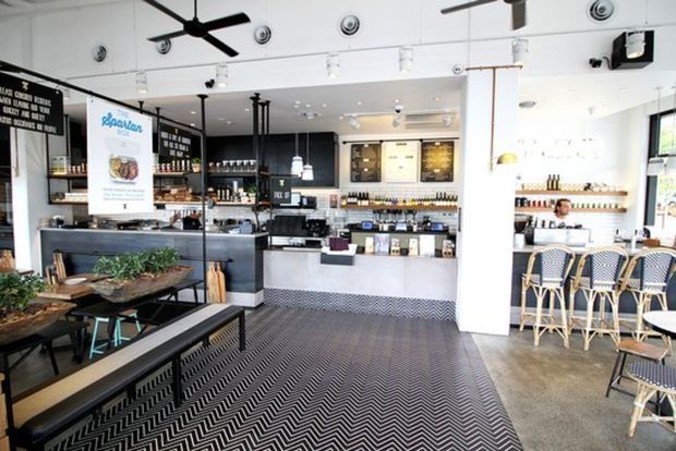 Bright, contemporary café interior with a service counter, espresso machine, pastry and beverage displays, bar seating, pendant lights, black ceiling fans, and a patterned black-and-white area rug over a polished concrete floor. The space combines white walls and columns with dark shelving, metal-framed communal furniture, and woven café chairs.