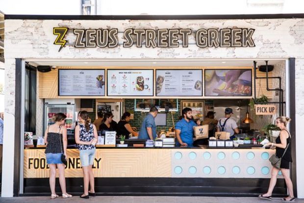 Front-facing view of a compact Greek street-food takeaway kiosk with a prominent Zeus Street Greek sign, illuminated menu boards, open service window, counter, staff, and customers. The fit-out combines light timber-patterned panels, pale blue circular accents, black trim, and a distressed white fascia.