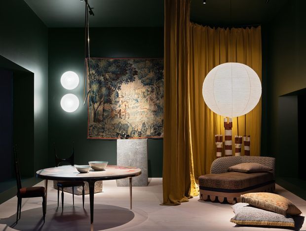 A moody, highly styled interior vignette with deep green walls, a round dining table and chairs, a large framed tapestry, sculptural globe lighting, and dramatic mustard-gold floor-to-ceiling curtains. A patterned upholstered chaise and floor cushions create a luxurious lounge corner, while contemporary lighting and vintage-inspired furniture add an eclectic, theatrical character.