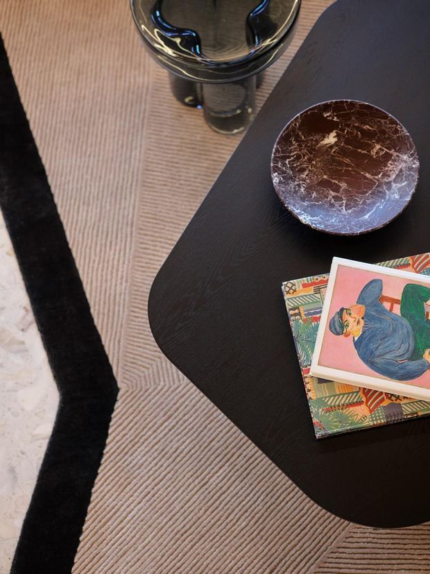 Close-up overhead view of a dark rounded-edge coffee table with a black wood-grain finish, positioned on a textured beige rug with a bold black border. The table holds a sculptural dark glass vessel, a marbled brown decorative bowl, and colourful art books. The composition has a refined, contemporary decorative styling.
