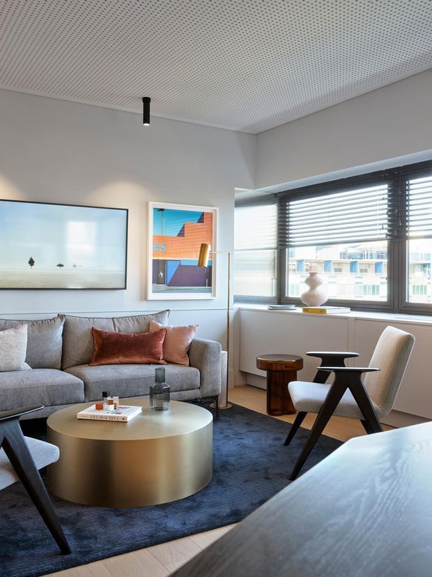 A stylish contemporary living room with a grey sectional sofa, round metallic coffee table, dark rug, sculptural lounge chairs, and framed artwork. Large windows with dark horizontal blinds provide abundant daylight and views of neighboring buildings. The room has a perforated acoustic ceiling, neutral grey walls, light timber flooring, and a refined modern fit-out.