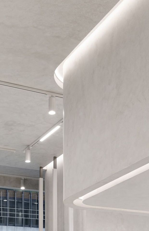 A close-up architectural interior view dominated by sculptural curved plaster or concrete walls and a softly rounded soffit with concealed cove lighting. The space has a pale monochromatic palette, exposed concrete-like ceiling surfaces, linear track lighting, cylindrical pendant spotlights, slender columns, and large gridded glazing in the background. The composition feels like a refined modern commercial or public interior.