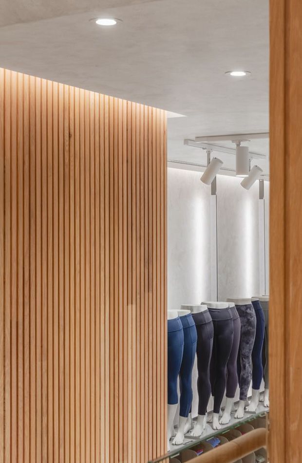 A minimalist retail clothing interior viewed through a narrow opening, featuring a full-height vertical timber slat partition in the foreground, warm wood framing, white textured walls, recessed ceiling lights, and track spotlights illuminating rows of mannequin lower bodies dressed in athletic leggings.