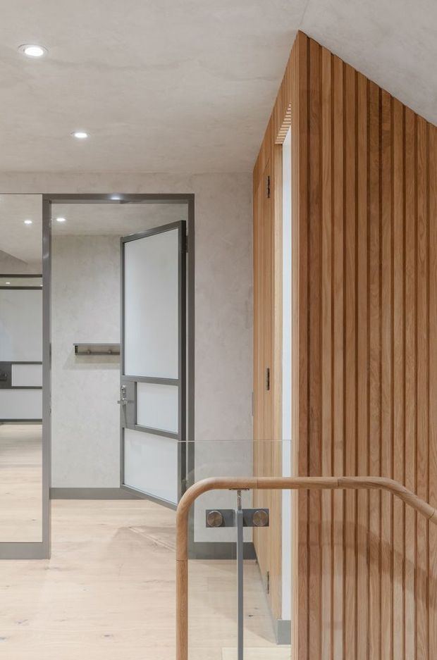 Interior view of a contemporary stair landing or hallway with a warm vertical timber slat feature wall, pale polished flooring and plastered ceiling. A glass balustrade with rounded timber handrail borders the stair, while a grey-framed glazed door and doorway lead to adjacent spaces.