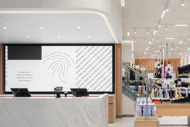 A bright contemporary retail clothing store interior featuring a large white reception or checkout counter in the foreground, integrated point-of-sale terminals, and a graphic feature wall with typographic artwork. Behind it, open retail displays, hanging clothing racks, glass balustrades, timber-toned joinery, and extensive ceiling track lighting create a clean, spacious commercial fit-out.