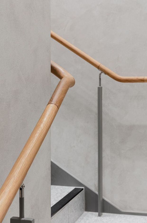 Close-up architectural photograph of a contemporary stairway with rounded natural timber handrails, slim dark metal supports, grey polished plaster or concrete walls, and pale stone or concrete stair treads. The composition focuses on the handrail detailing and stair geometry.