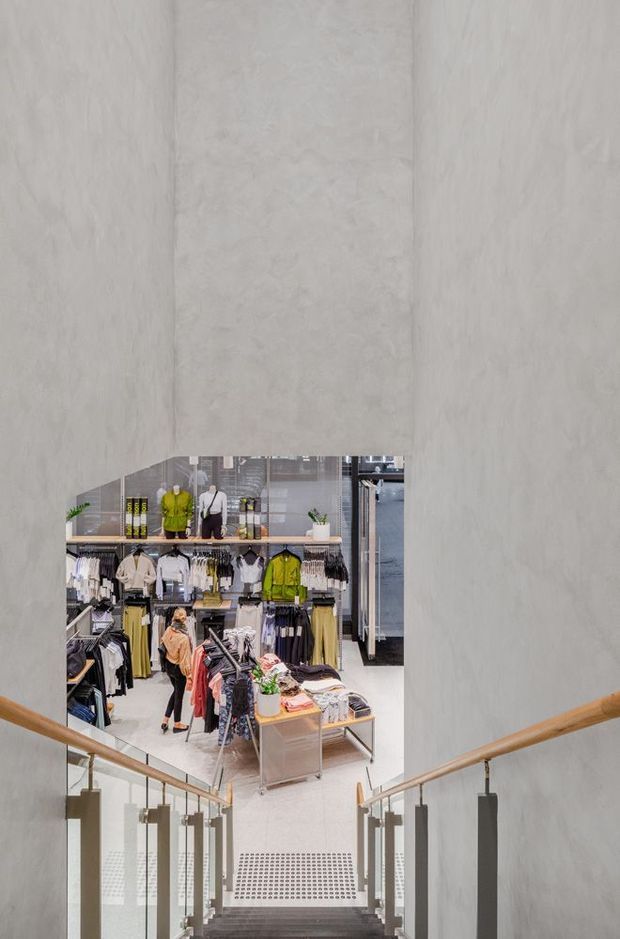A view down a modern stairway with glass balustrades and timber handrails, looking into a bright multi-level clothing retail store. The retail space has polished light flooring, wall-mounted clothing displays, racks, mannequins, shelving, and a large curved opening framed by pale textured walls.
