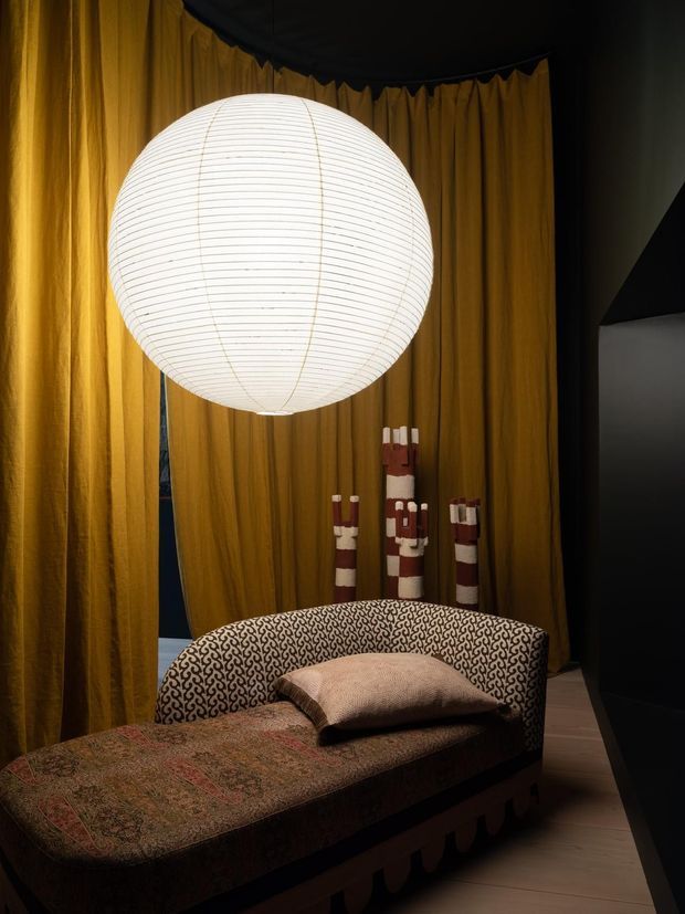 A moody, theatrical interior vignette featuring a curved upholstered chaise or daybed in a dark patterned fabric, positioned beneath a large glowing spherical paper pendant light. Mustard-gold drapery lines the background, with sculptural red-and-cream decorative objects behind the seating. The space has a richly layered, eclectic feel with dramatic lighting and dark surrounding surfaces.