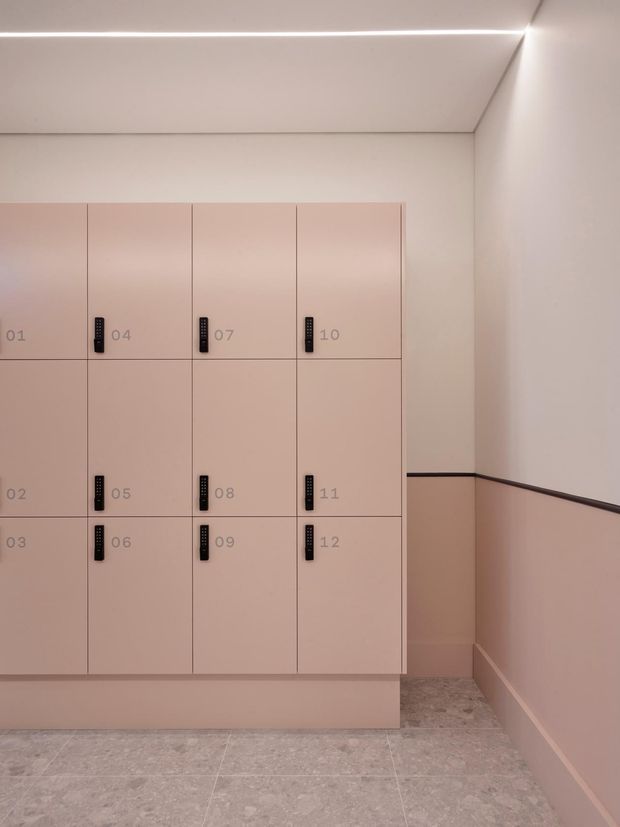 A minimalist locker area with a wall-mounted grid of pale pink-beige lockers numbered 01–12, each fitted with black combination locks. The surrounding walls and trim are painted in matching soft blush tones, with a dark horizontal rail and a light grey stone-look tiled floor. A slim recessed linear light runs along the ceiling edge.