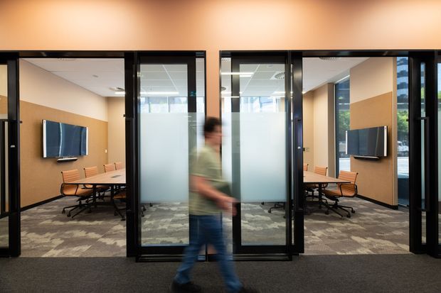 A contemporary commercial office interior viewed through black-framed glazed partitions. The image shows meeting rooms with timber-toned wall panels, mounted screens, conference tables, office chairs, frosted glass sections, and patterned carpet. A blurred person walks across the foreground.