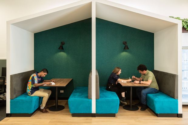 A bright contemporary commercial interior with two semi-enclosed booth seating areas. Each booth has a deep teal upholstered bench, dark teal textured feature wall, white geometric surround, small wall sconce, and a dark wood table with black pedestal base. People are seated working or conversing, suggesting a café, coworking space, or informal meeting area.